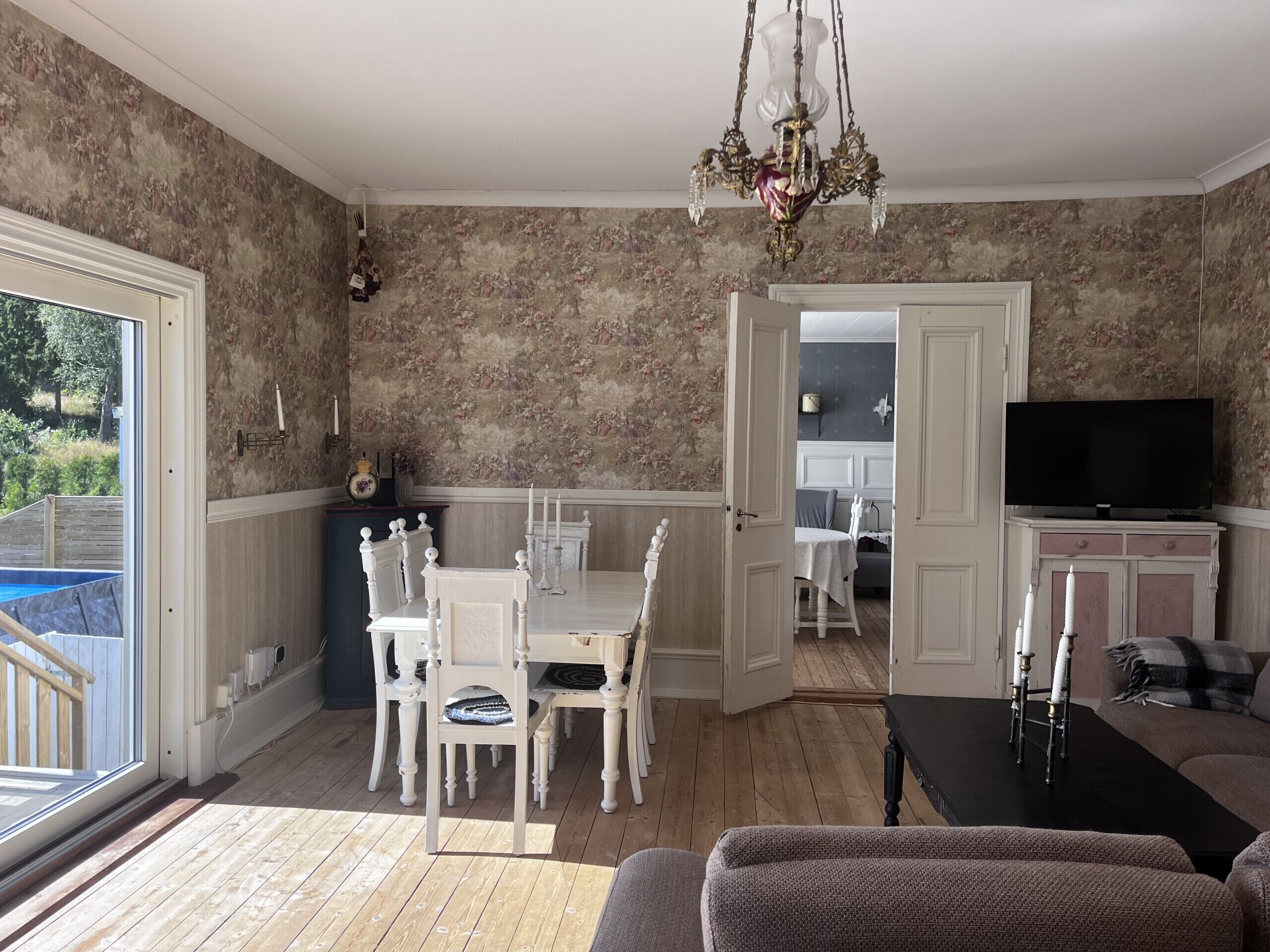 Un coin repas avec une table et des chaises, un lustre et un téléviseur, dans une pièce dont les murs sont recouverts d’un papier peint à motifs floraux.