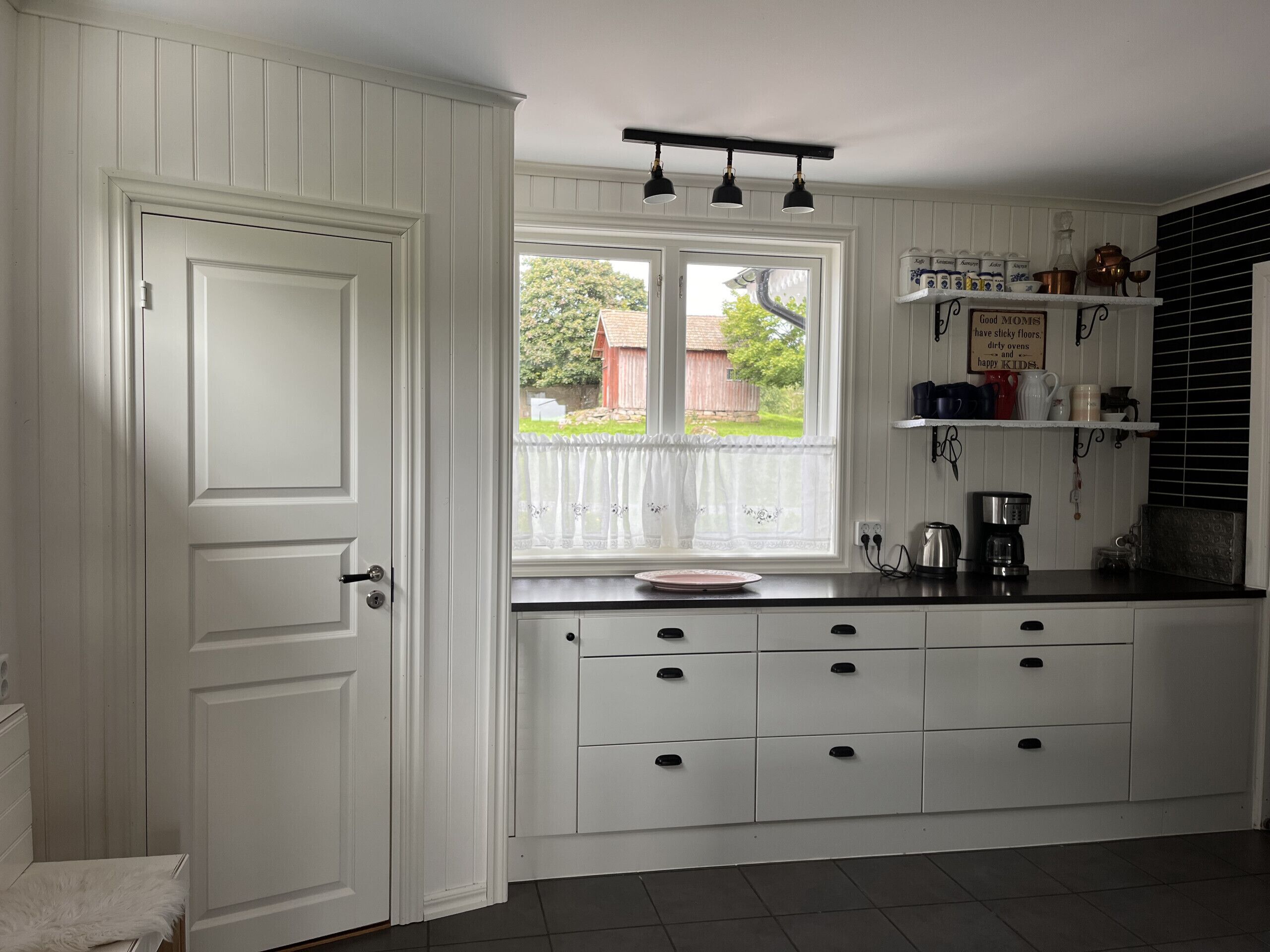 A kitchen with white cabinetry, a window with a view of the outside, and a shelf with kitchen items.
