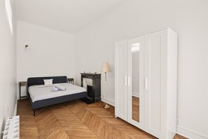 Ein Schlafzimmer mit Bett, Nachttisch, Lampe, Kleiderschrank und einem Parkettboden mit Fischgrätenmuster.