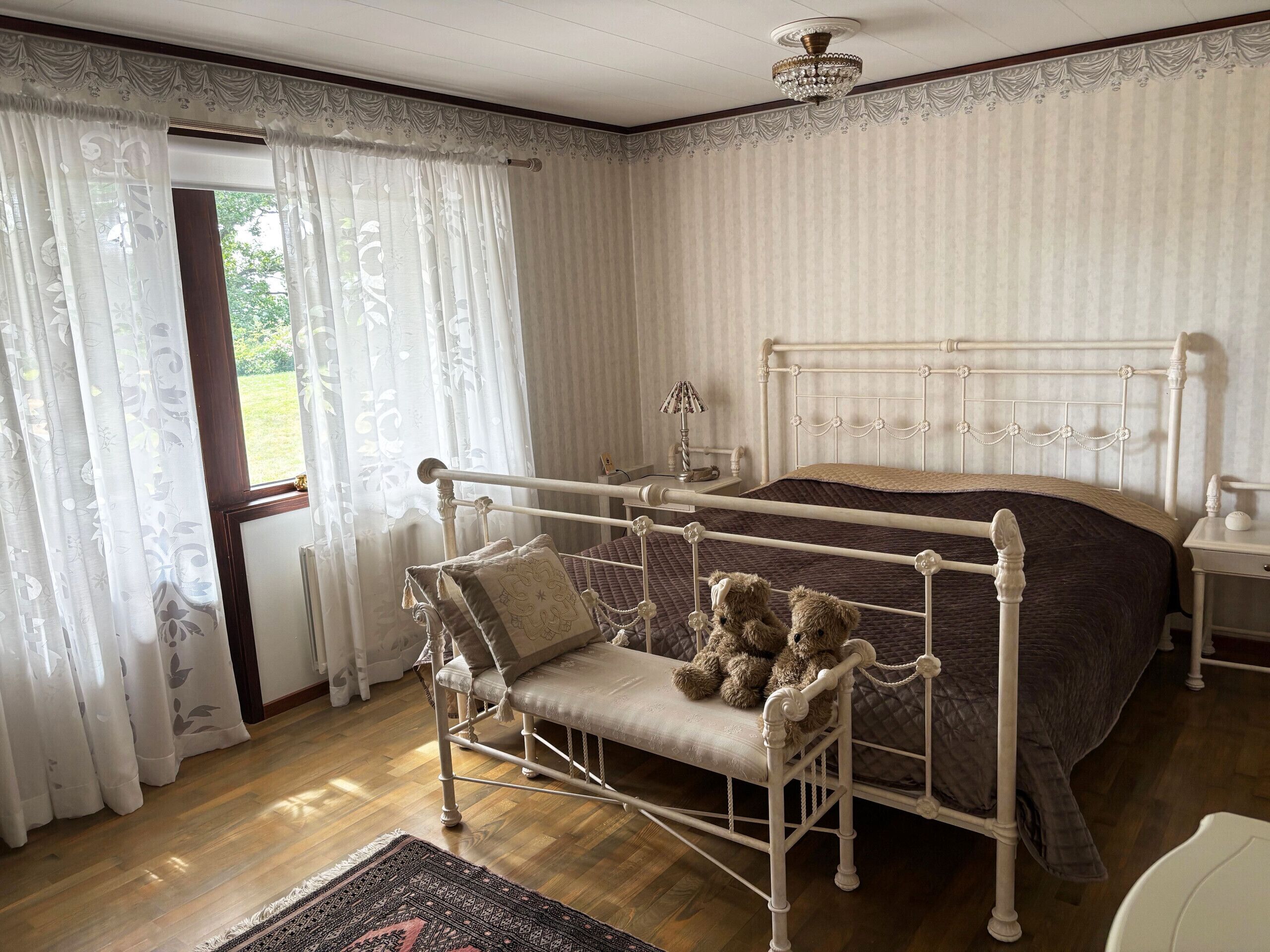 A bedroom with a metal bed frame, a bench with pillows, a bedside table with a lamp, and a view through a window with curtains.