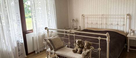 A bedroom with a metal bed frame, a bench with pillows, a bedside table with a lamp, and a view through a window with curtains.