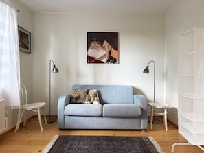 Un salon avec un canapé bleu clair, deux tables d’appoint, un tapis à motifs et un tableau accroché au mur.