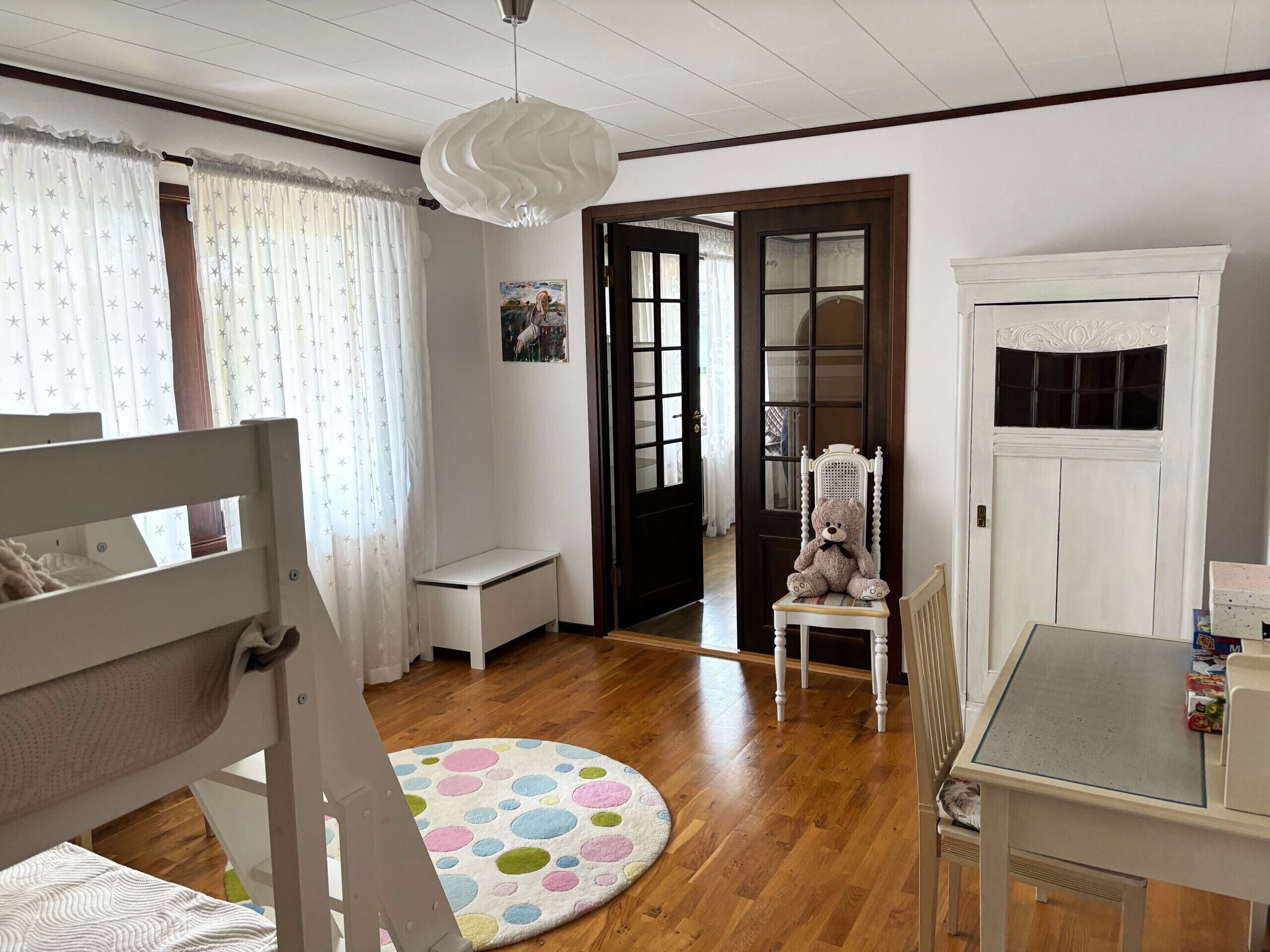 A room with a wooden floor, a round rug with colorful circles, a white desk, a chair, a teddy bear on a chair, and a white door with glass panels.