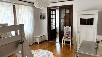 A room with a wooden floor, a round rug with colorful circles, a white desk, a chair, a teddy bear on a chair, and a white door with glass panels.