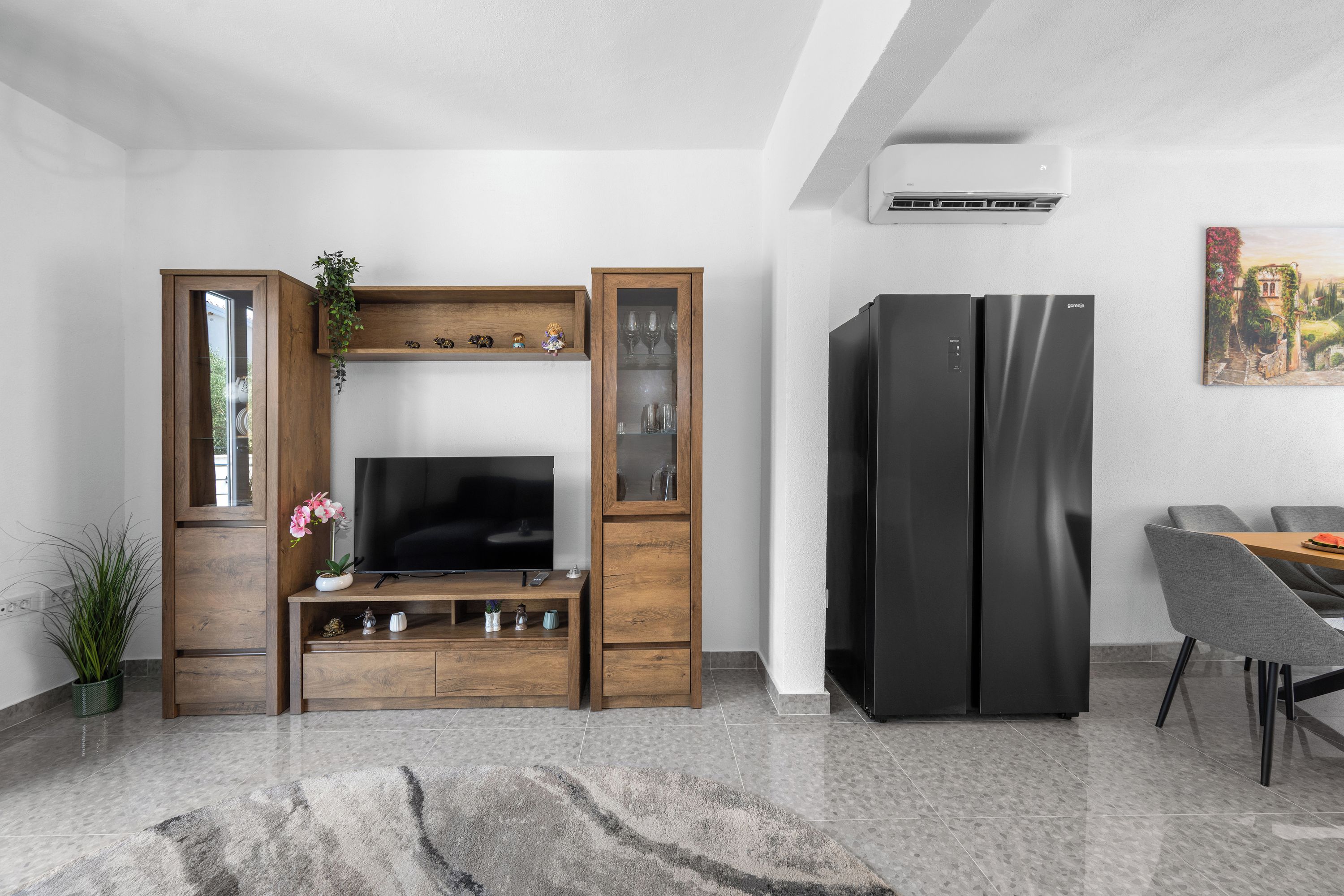 Una sala moderna con televisor de pantalla plana, mueble de entretenimiento de madera, dos refrigeradores negros y un comedor con sillas grises.