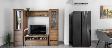 A modern living room with a flat-screen TV, wooden entertainment unit, two black refrigerators, and a dining area with grey chairs.
