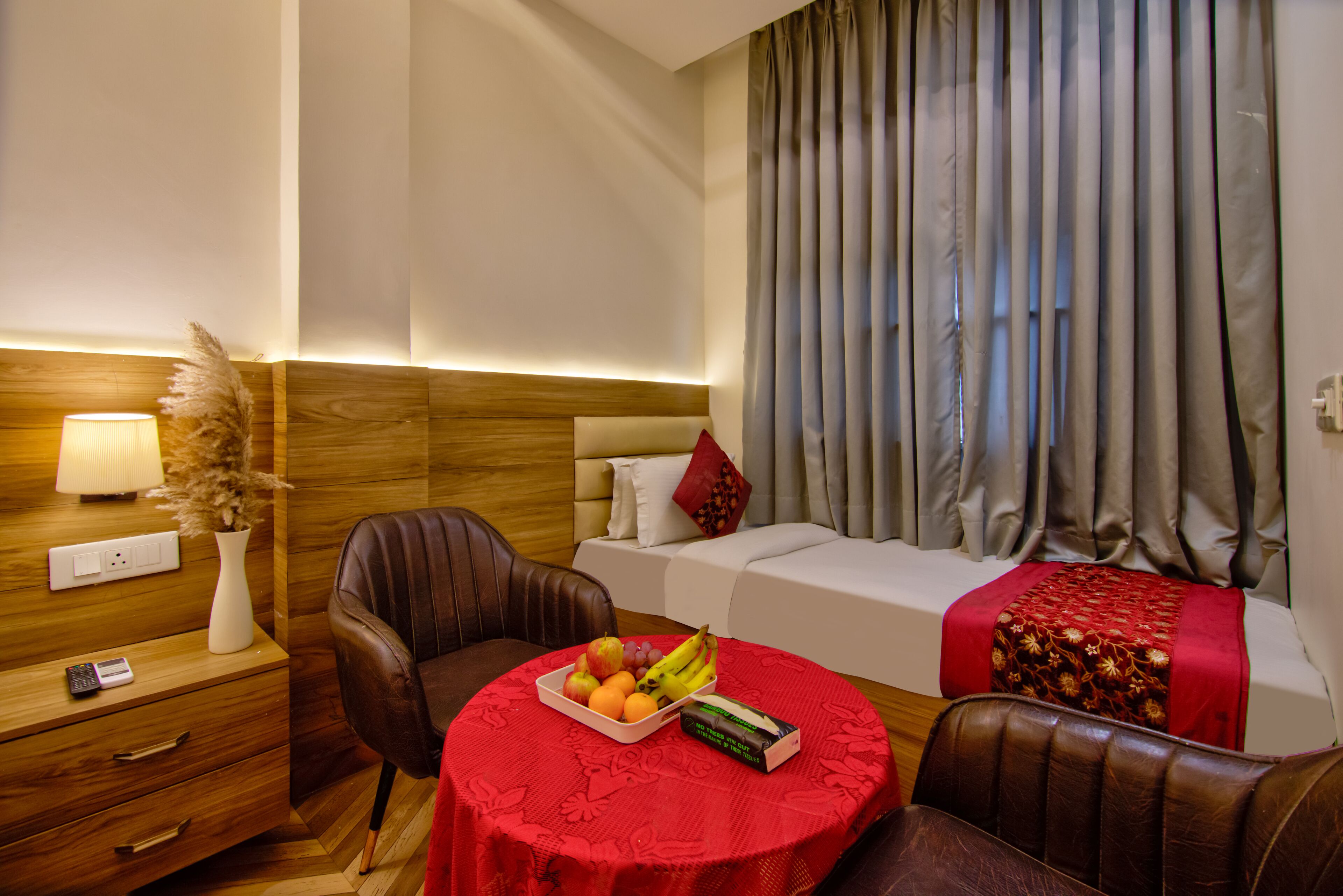 Quarto de hotel com cama, escrivaninha com abajur, cadeira, mesa com frutas e janela com cortinas.
