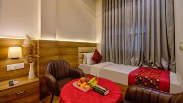 Quarto de hotel com cama, escrivaninha com abajur, cadeira, mesa com frutas e janela com cortinas.