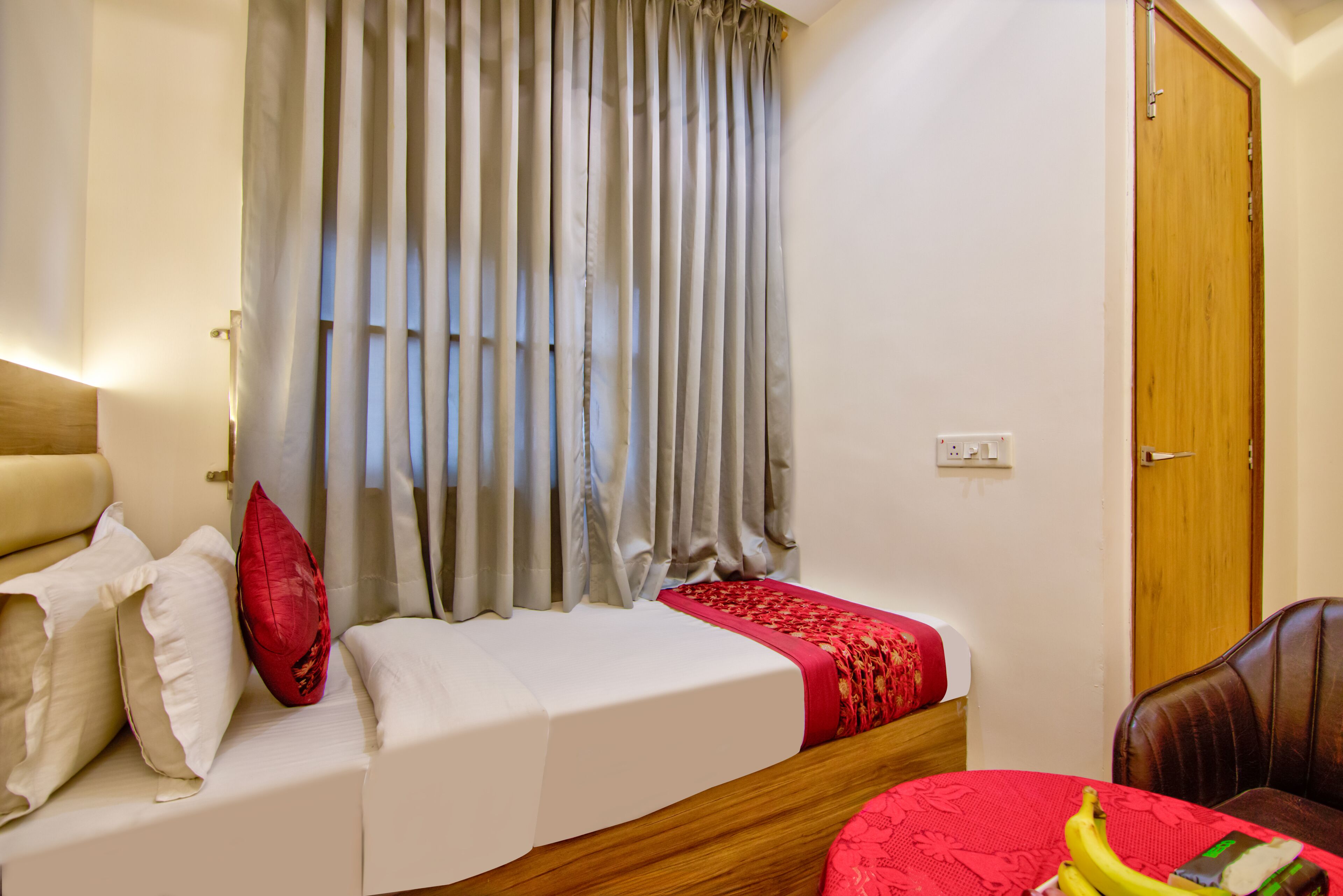 A hotel room with a bed, a wooden headboard, a red and white bedspread, a red pillow, a window with grey curtains, and a wooden door.