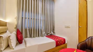 Une chambre d’hôtel avec un lit, une tête de lit en bois, une couvre-lit rouge et blanche, un oreiller rouge, une fenêtre avec des rideaux gris et une porte en bois.