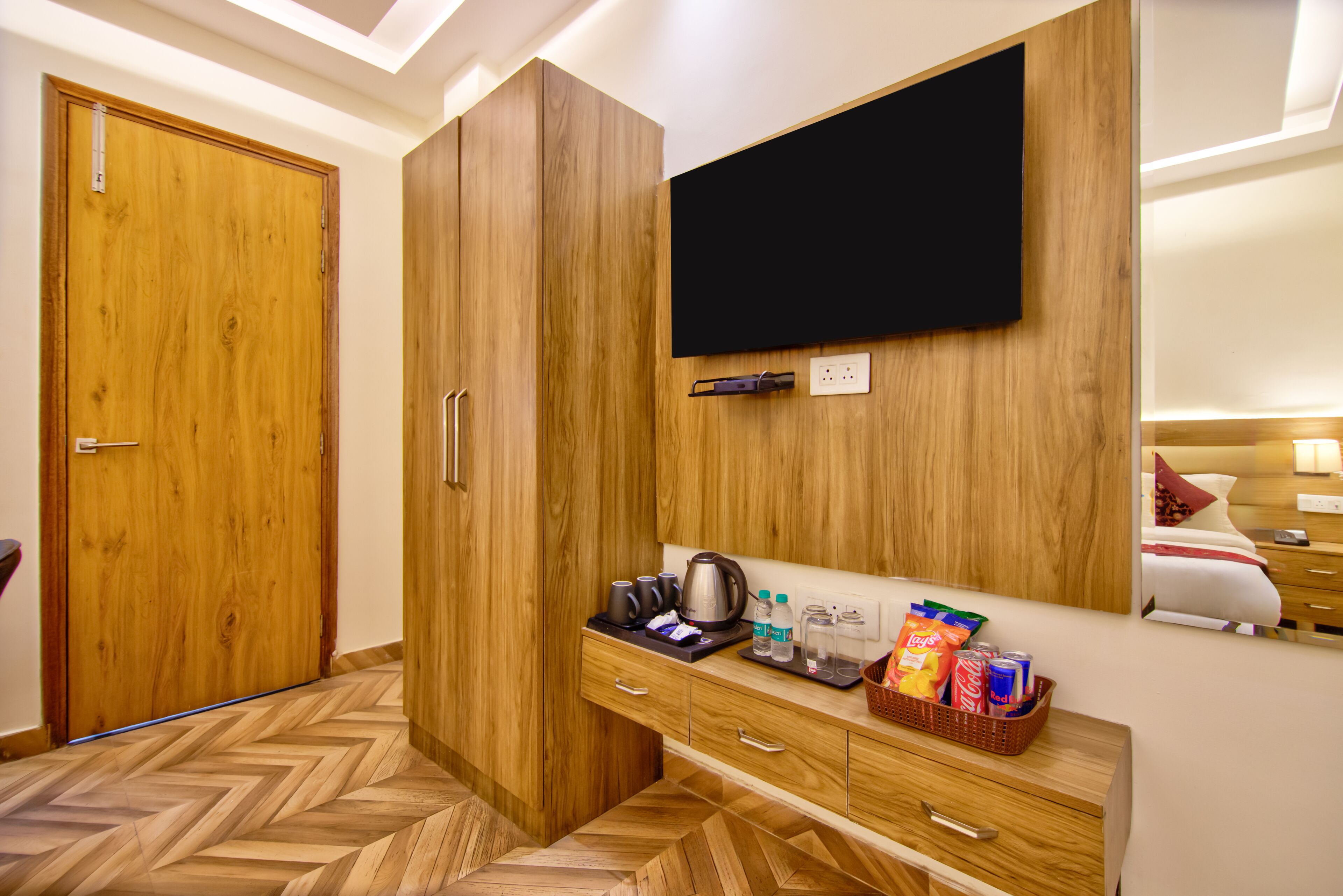 Quarto de hotel com guarda-roupa de madeira, televisão de ecrã plano na parede, kitchenette compacta com chaleira e minibar com snacks e bebidas.