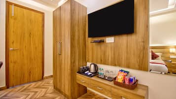 Quarto de hotel com guarda-roupa de madeira, televisão de ecrã plano na parede, kitchenette compacta com chaleira e minibar com snacks e bebidas.