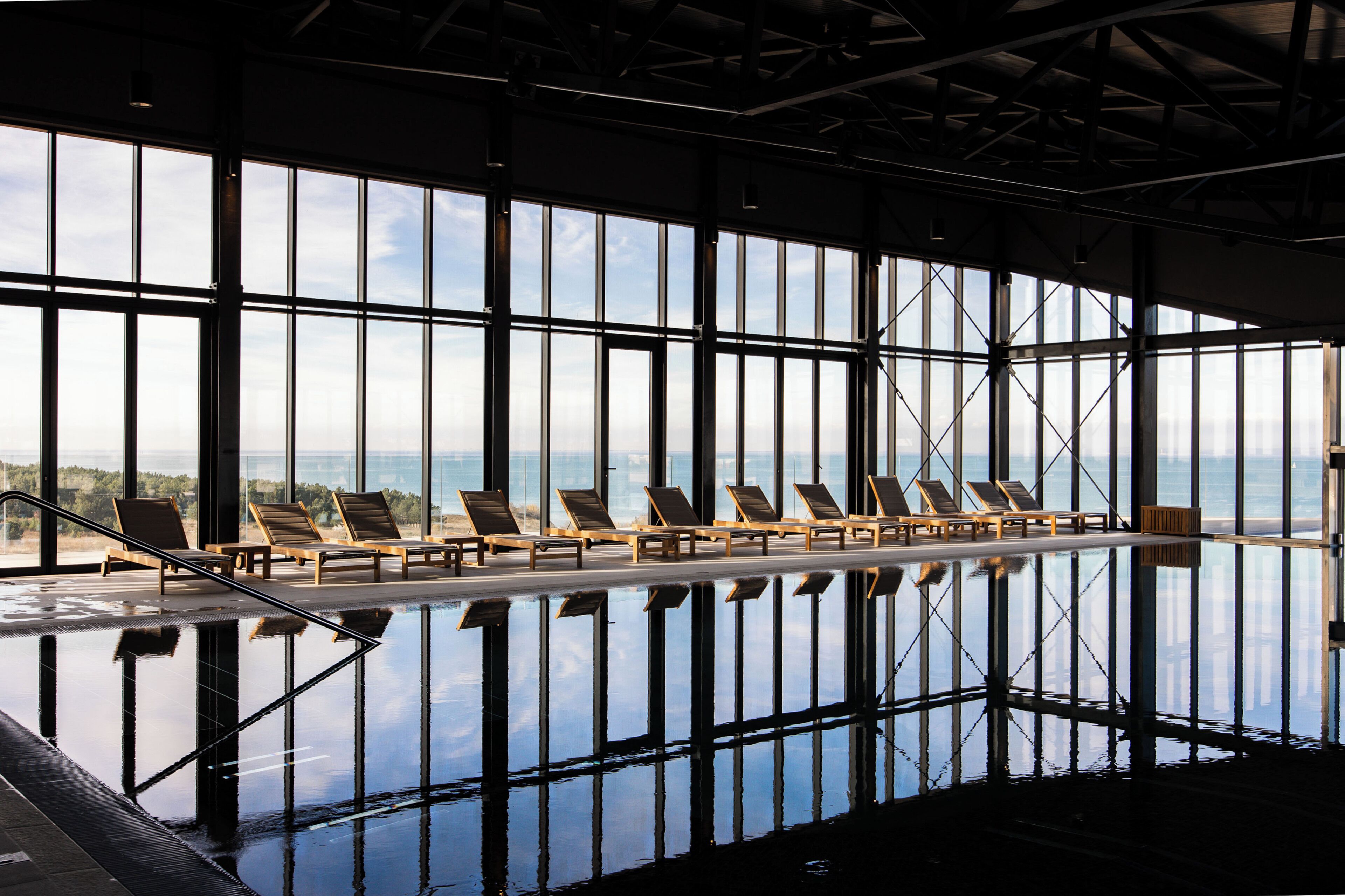 Ein großer, überdachter Swimmingpool mit Glaswänden, mehreren Liegestühlen und Blick auf den Ozean.