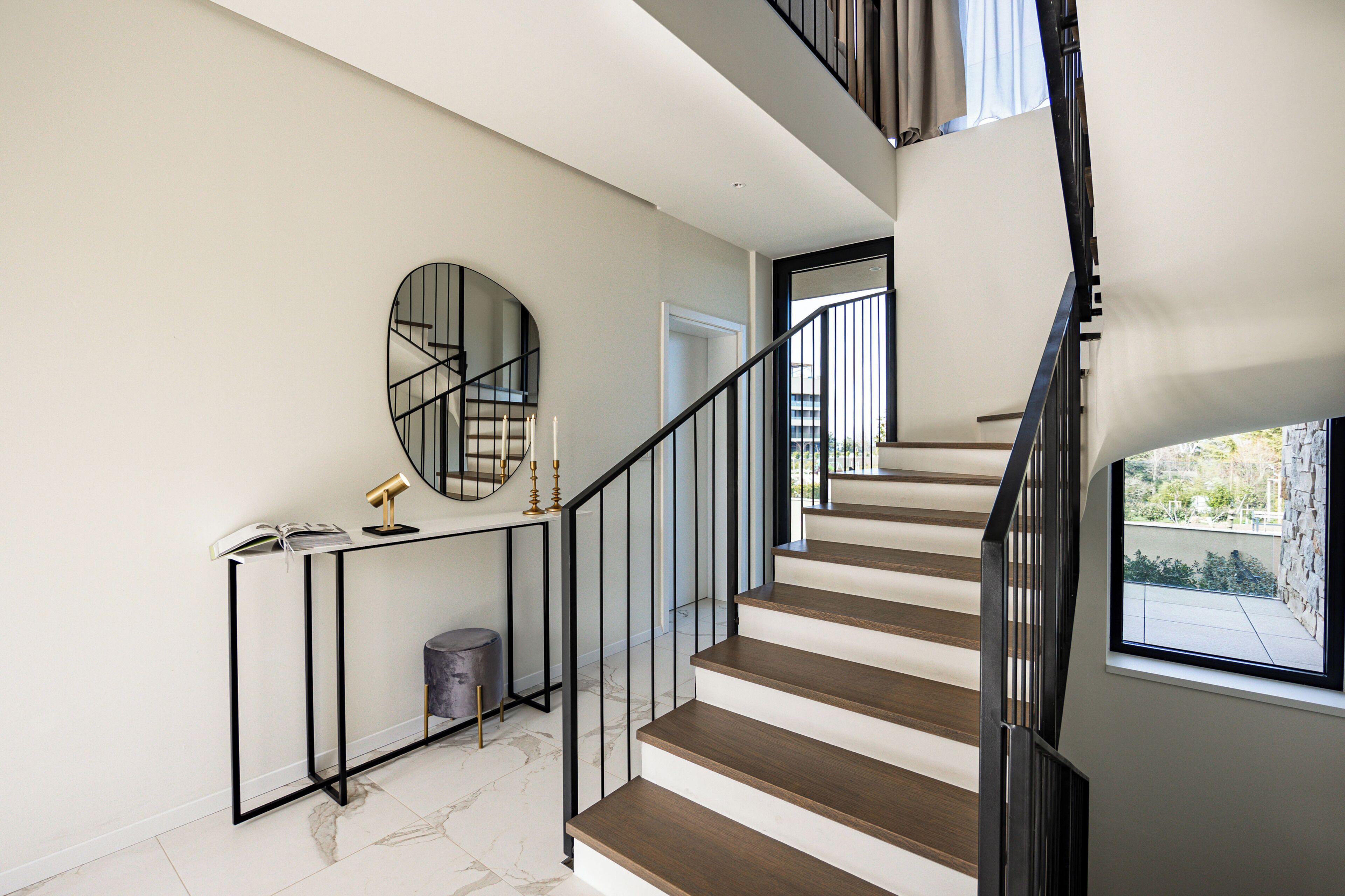 Un escalier moderne avec une rampe noire, un miroir rond accroché au mur et une petite table sur laquelle se trouvent une lampe et des bougies.