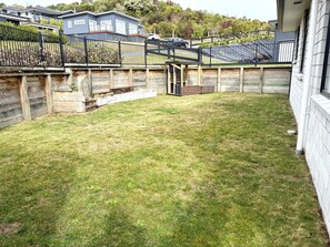 Un jardin à l’arrière d’une maison, avec une terrasse en bois, une zone délimitée par une clôture et la maison en arrière-plan.