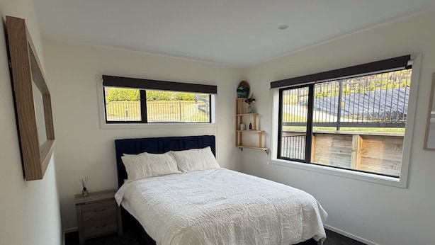 A bedroom with a bed, a nightstand, a window with a view of a fence, and a shelf with decorative items. - The Lakeview Residence (Kinloch)
