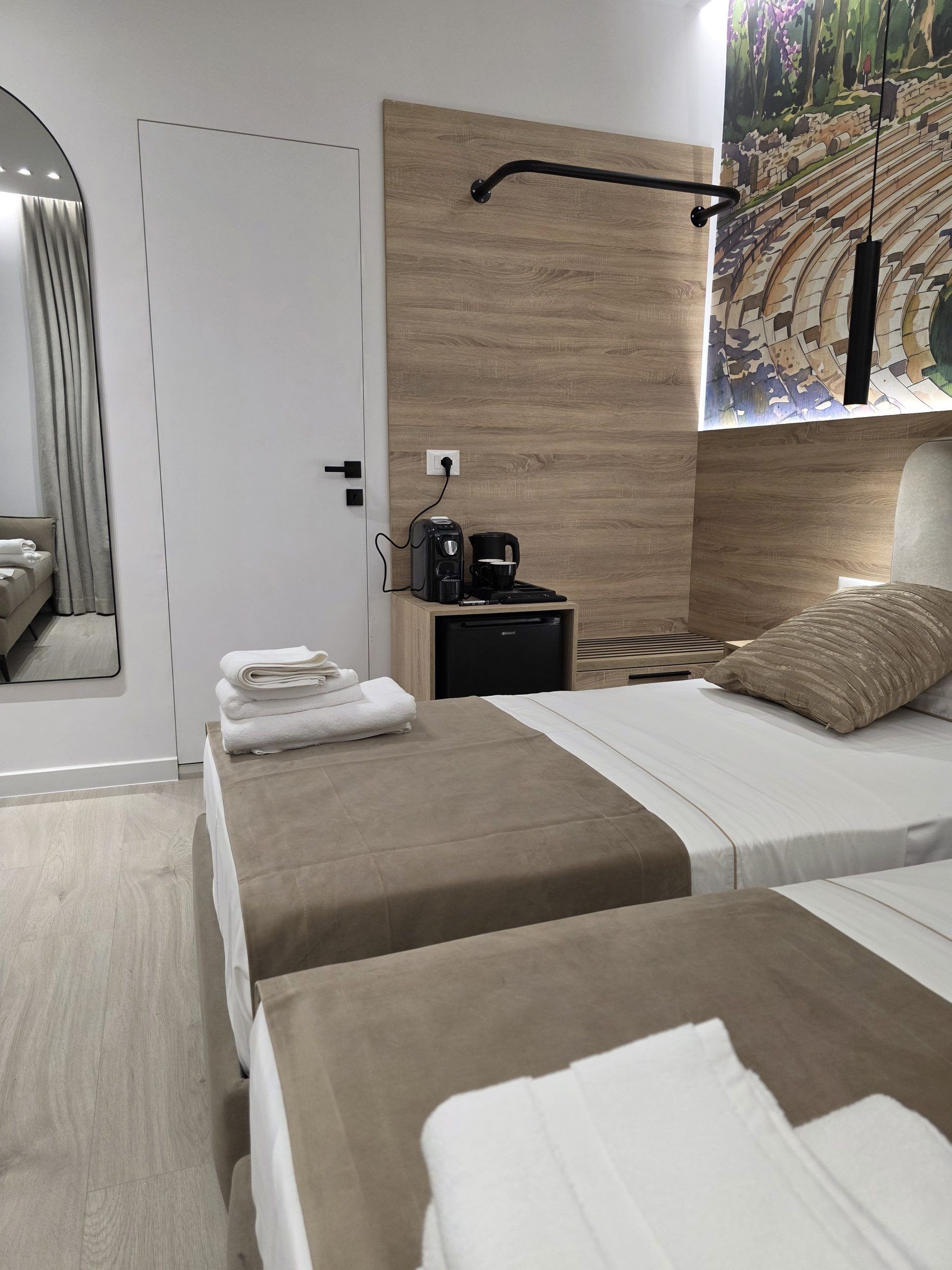A hotel room with a bed, a nightstand with a coffee maker, a mirror, and a painting on the wall.