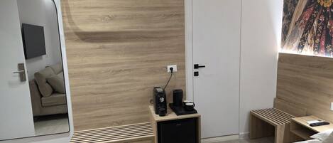 A modern bedroom with a wooden headboard, a bedside table with a coffee maker, a small cabinet, and a bed with white linens.