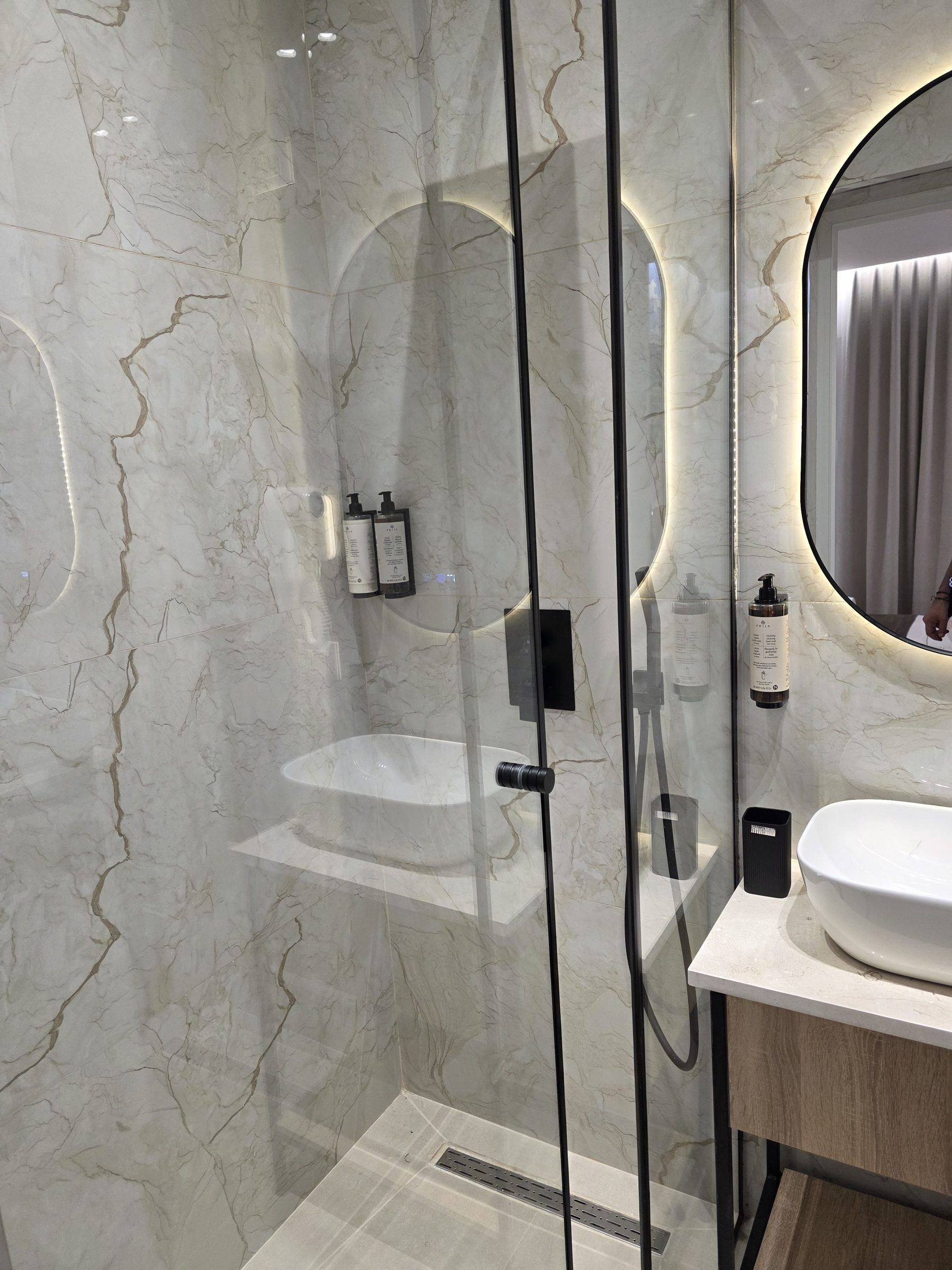 Un baño moderno con ducha envuelta en vidrio, lavabo blanco y pared de mármol.