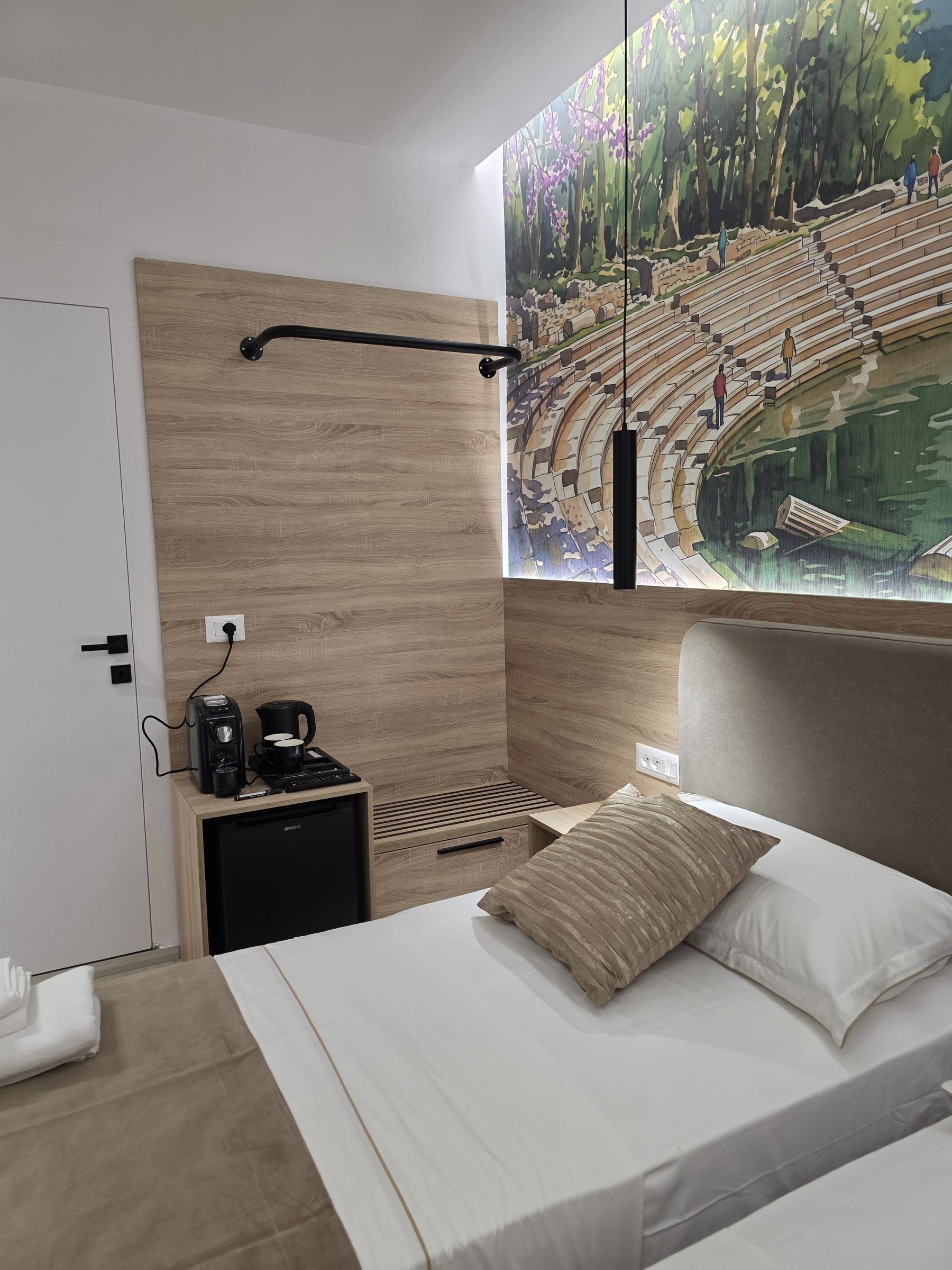 Habitación de hotel moderna con cama, mesita de noche con cafetera, televisor montado en la pared y ventana con vista a un parque.