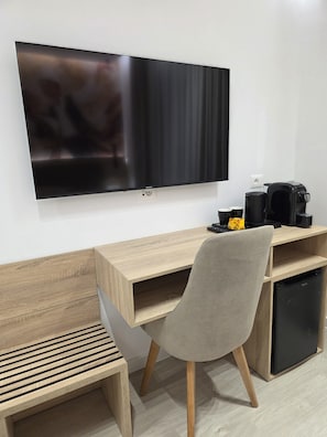 A modern room with a flat-screen TV mounted on the wall, a wooden desk with a chair, a coffee maker, and a small refrigerator. - The Story Hotel (Tirana)