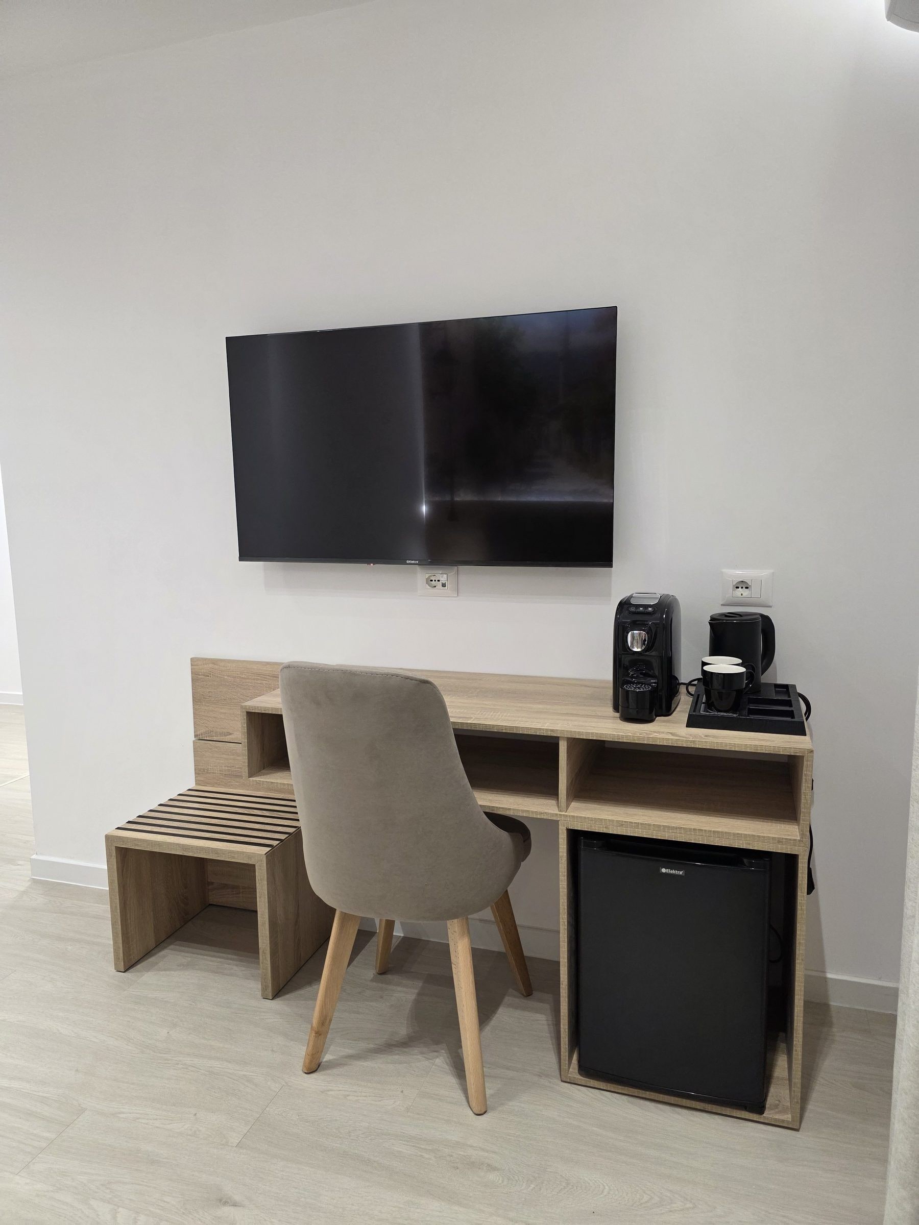 Un espacio de trabajo moderno con una televisión de pantalla plana, un escritorio, una silla, una cafetera y un refrigerador pequeño.