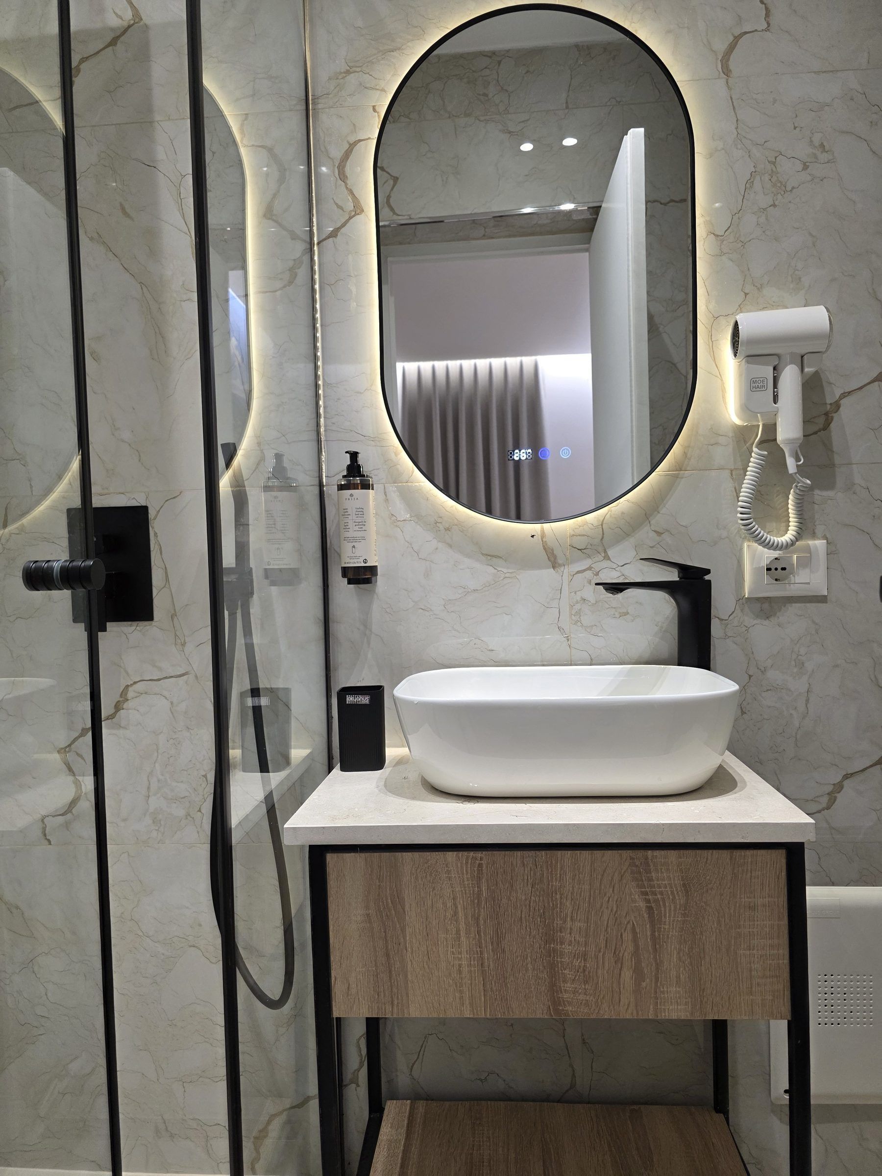 Une salle de bain moderne avec un mur en marbre, un miroir rond, un lavabo blanc et un sèche-cheveux.