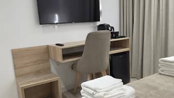 Una habitación de hotel con una cama, un escritorio con una silla, un televisor montado en la pared, una cafetera y una pila de toallas.