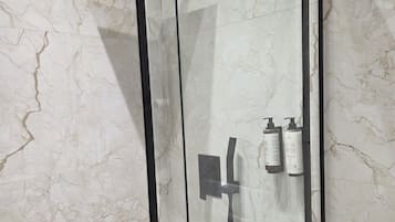 Ducha con paredes de vidrio, un revestimiento de mármol, cabezal de ducha manual y dos botellas de champú.
