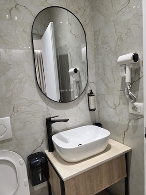 A bathroom with a marble wall, a white sink, a black faucet, a round mirror, and a hairdryer mounted on the wall. - The Story Hotel (Tirana)