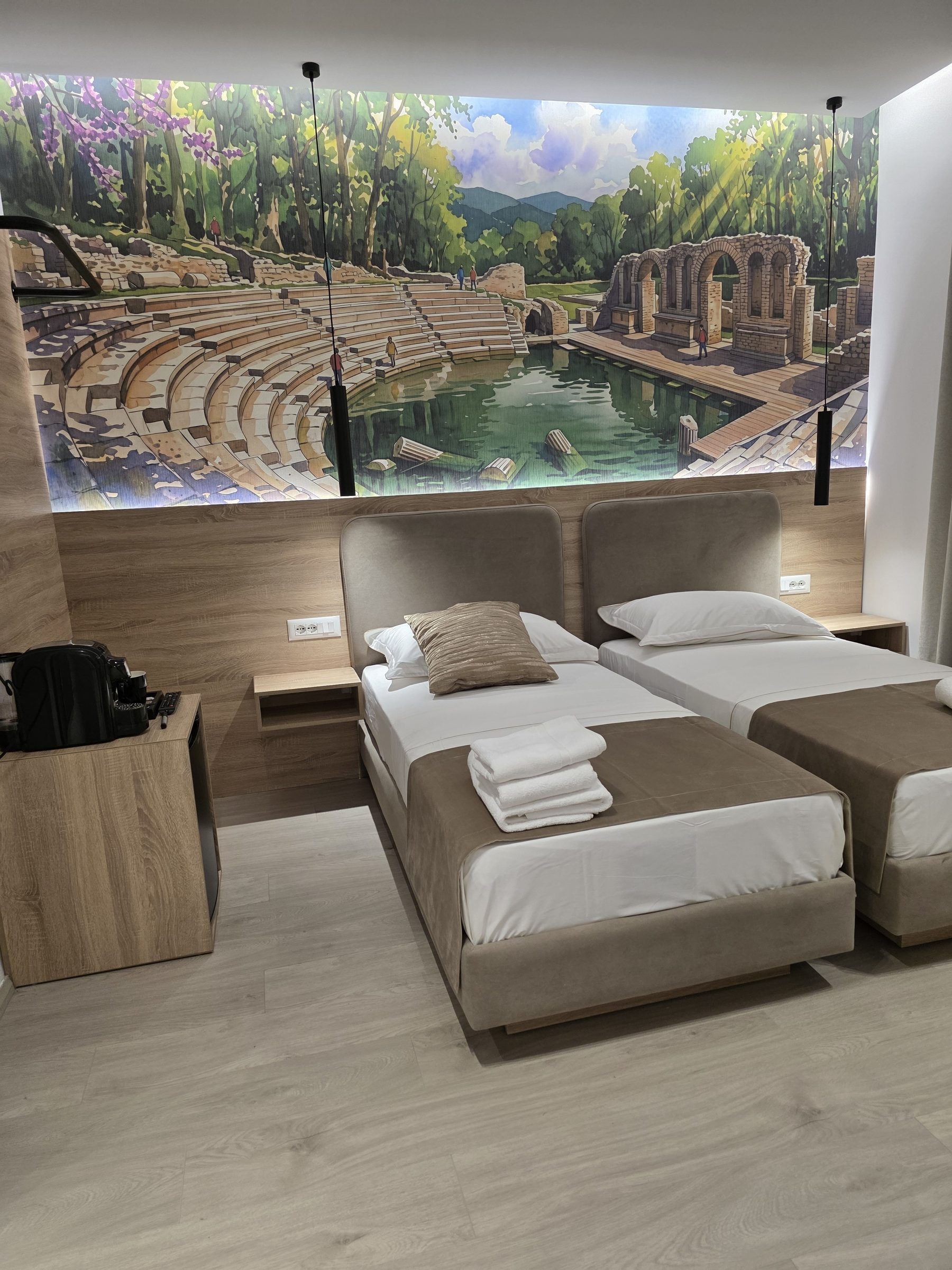 A hotel room with a large mural of a forest and ruins, two beds with white linens, a wooden side table, and a coffee machine.