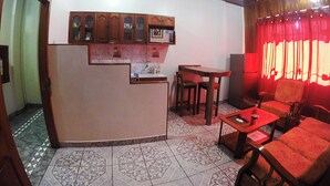 Una cocina con mostrador integrado, armarios de madera y suelo de baldosas. Hay una zona de comedor con una mesa y sillas, y un sofá rojo.