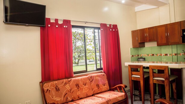 Un espacio habitable compacto con un sofá, un televisor montado en la pared, un ventanal con cortinas rojas y una kitchenette con armarios de madera.