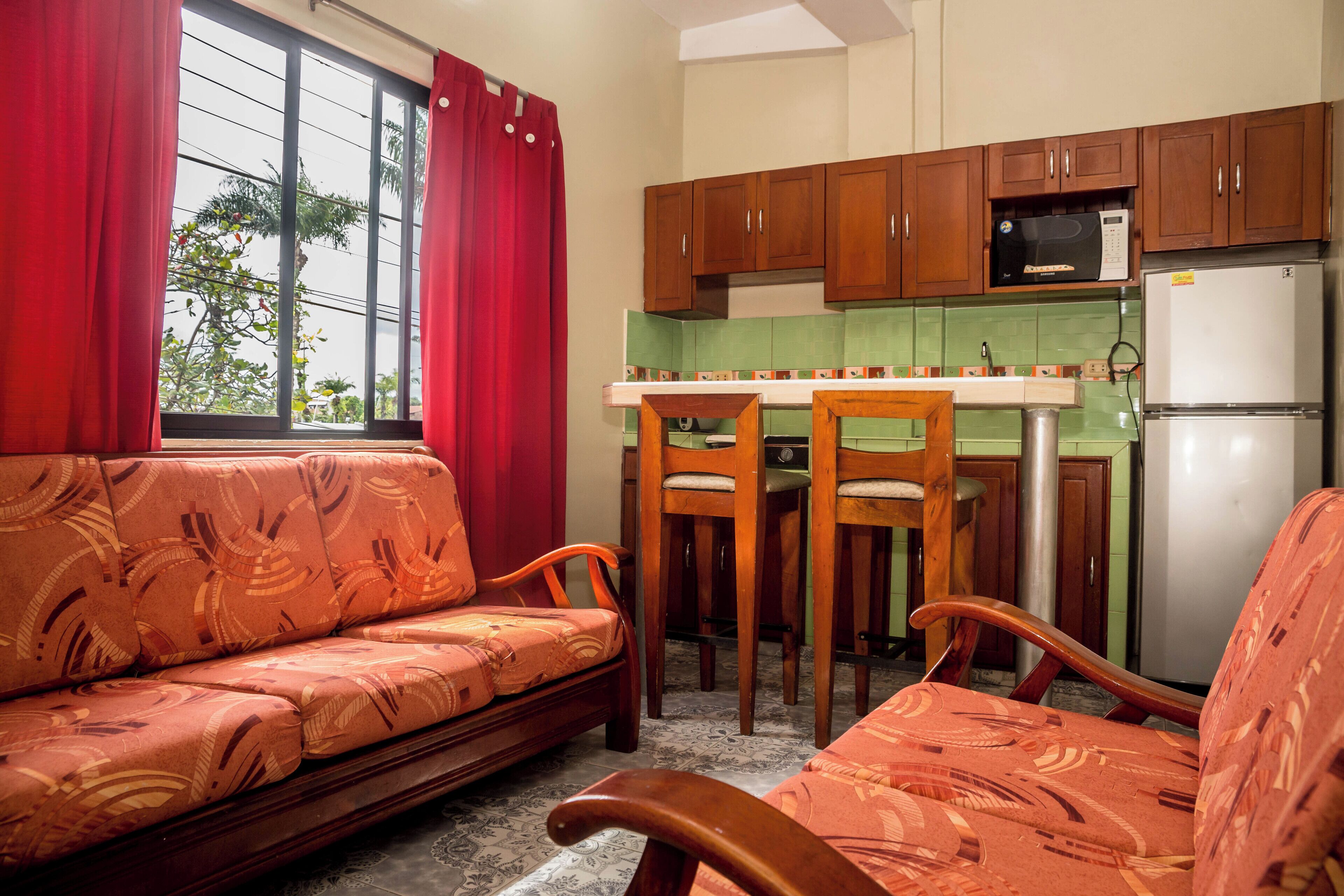 A compact living space with a sofa, chairs, a small kitchen area, and a window with curtains.