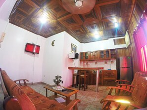 A living room with a wooden ceiling, a television mounted on the wall, a kitchen area with wooden cabinets, and a dining table with chairs.