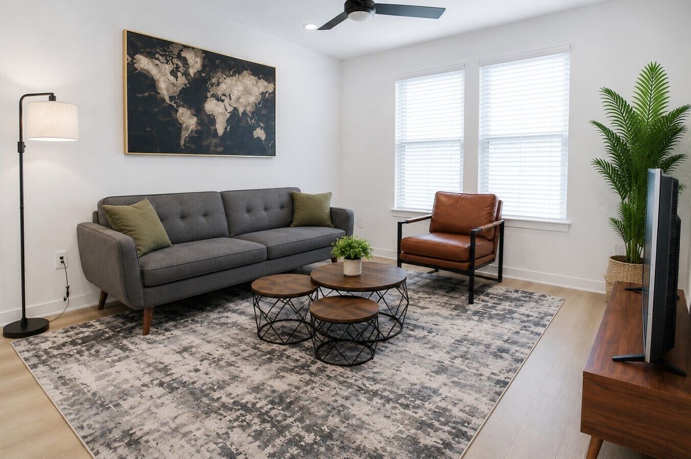 A living room with a gray sofa, a brown leather chair, a round coffee table, a floor lamp, a world map artwork, and a large window with blinds.