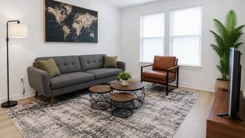 A living room with a gray sofa, a brown leather chair, a round coffee table, a floor lamp, a world map artwork, and a large window with blinds.