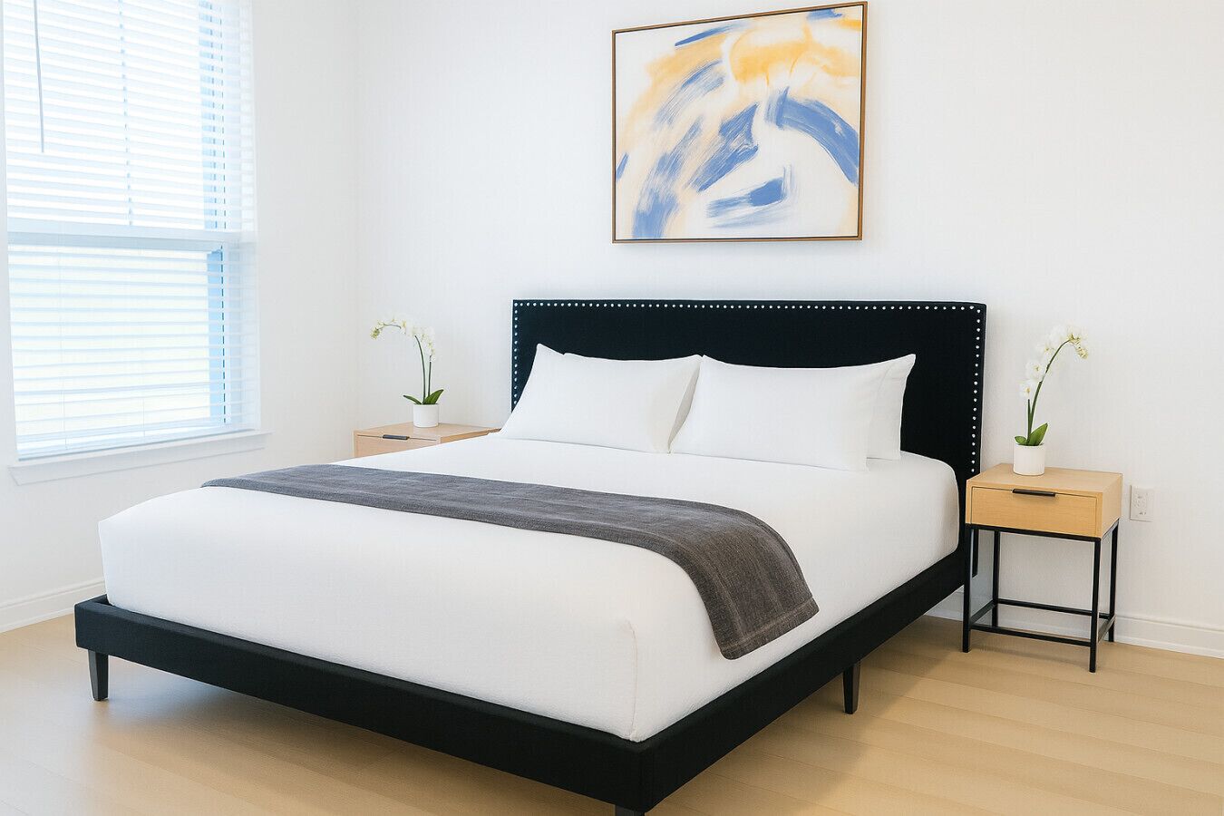 A modern bedroom with a black-framed bed, white bedding, and a gray throw blanket. There are two nightstands with white orchids, and a large abstract painting above the bed.