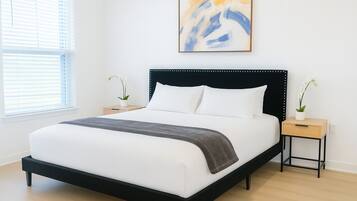 A modern bedroom with a black-framed bed, white bedding, and a gray throw blanket. There are two nightstands with white orchids, and a large abstract painting above the bed.