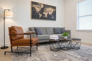 Una sala moderna con un sillón de cuero, sofá gris, una obra de arte con un mapa mundial, una lámpara de pie y una pequeña mesa con una planta.