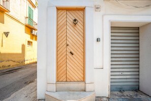 A building entrance with a wooden door featuring a chevron pattern, a metal doorbell, and a metal garage door to the right. - Domus Fisculi A: Authentic Apulian Stone-Vaulted Home (Fasano)