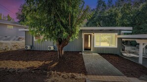 Una casa moderna amb un arbre al jardí, un porxo cobert i un camí que condueix a l'entrada.
