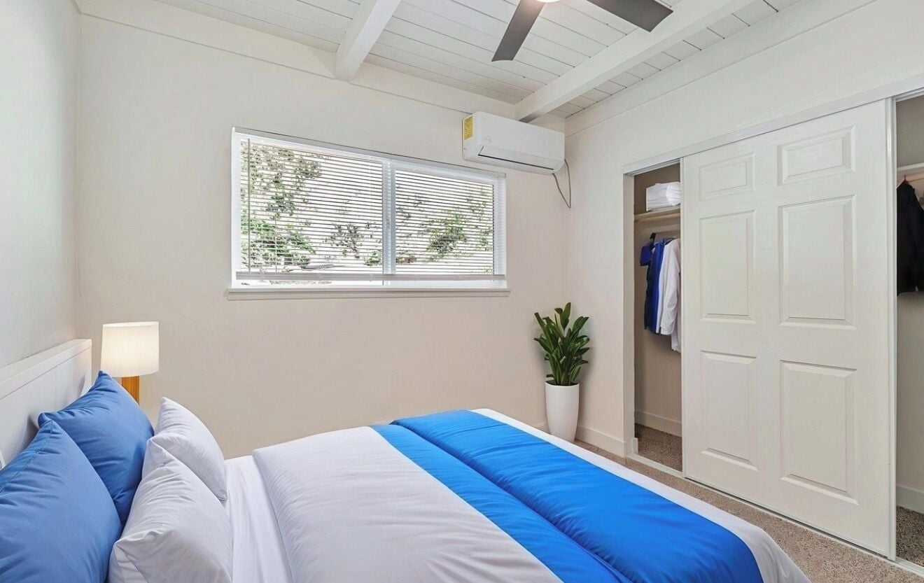 Un dormitorio con cama, ventana con persianas, unidad de aire acondicionado, armario y planta en maceta.
