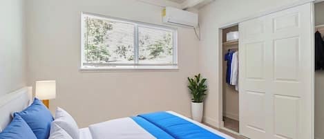 Un dormitorio con cama, ventana con persianas, unidad de aire acondicionado, armario y planta en maceta.