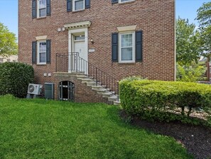 A two-story brick building with a white door, black shutters, and a small front porch with a metal railing. - 2 Mins from King St < > A Boutique Old Town Xcape (Alexandria)