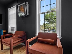 A room with two brown chairs, a window with a view of trees and parked cars, and a wall-mounted air conditioning unit. - 2 Mins from King St < > A Boutique Old Town Xcape (Alexandria)