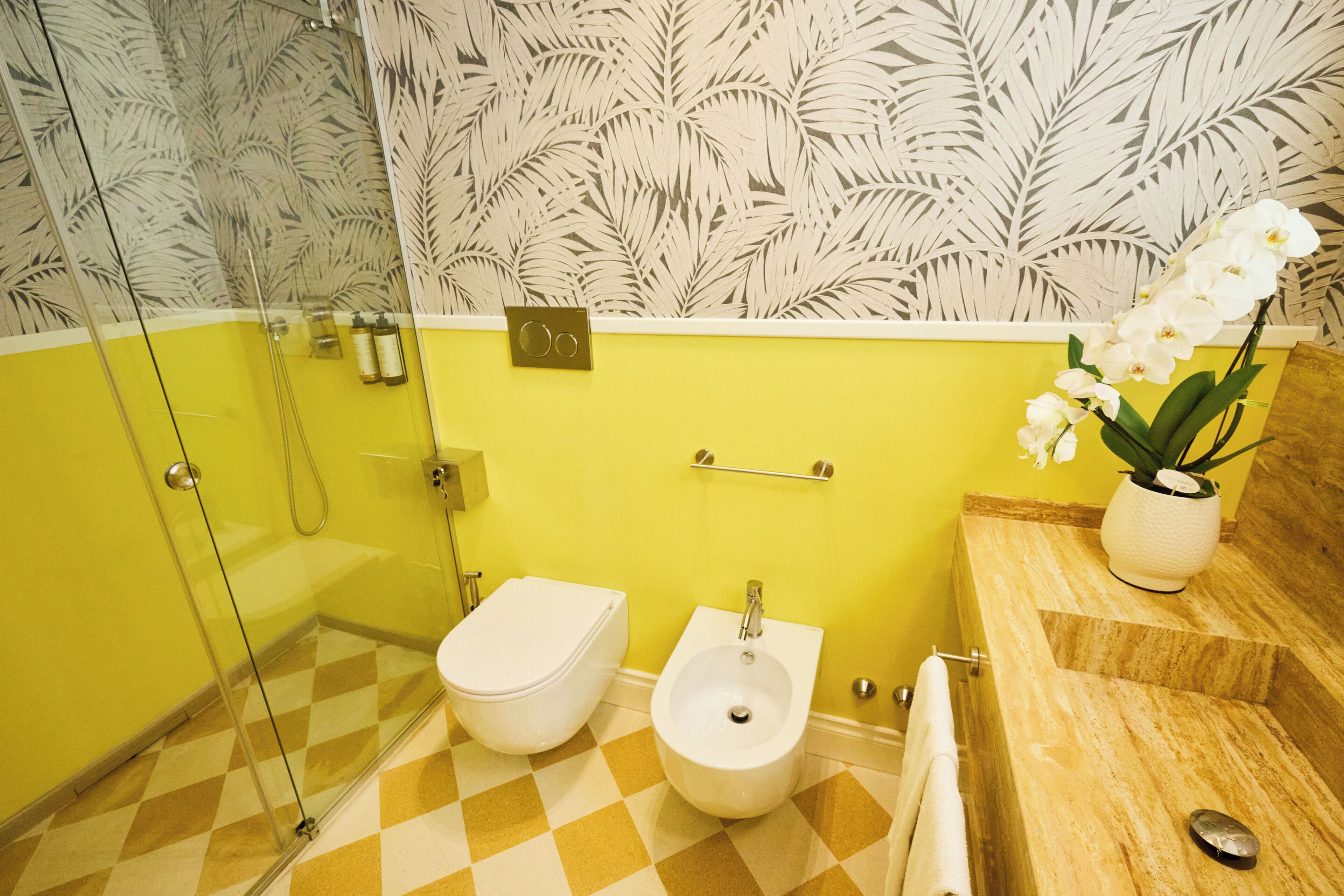 A bathroom with a checkered floor, a toilet, a bidet, a shower, and a sink with a potted orchid.