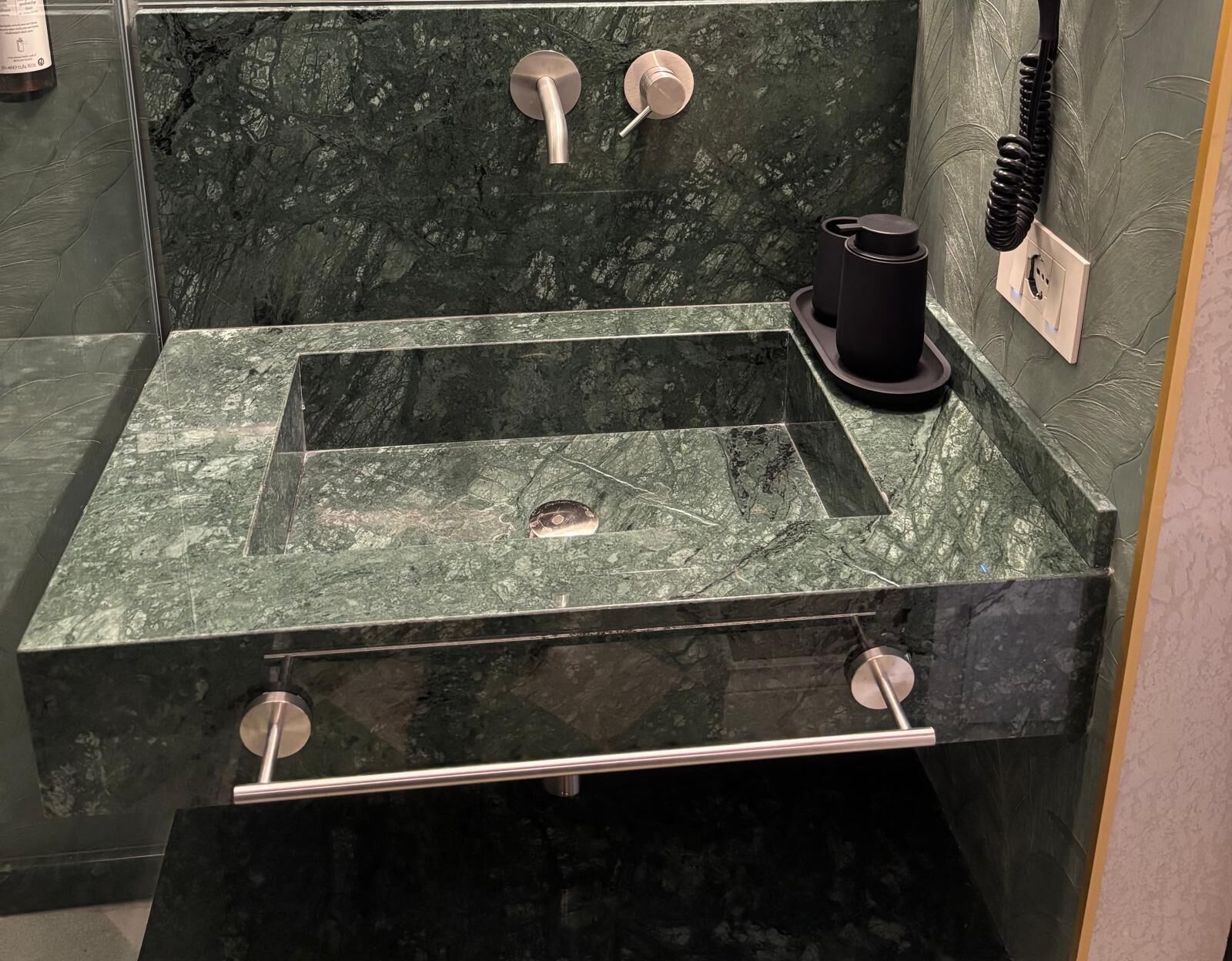 Un baño con un lavamanos rectangular, dos grifos, un dispensador de jabón negro y un perchero para toallas.