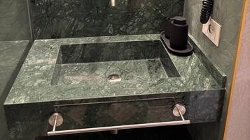 Un baño con un lavamanos rectangular, dos grifos, un dispensador de jabón negro y un perchero para toallas.