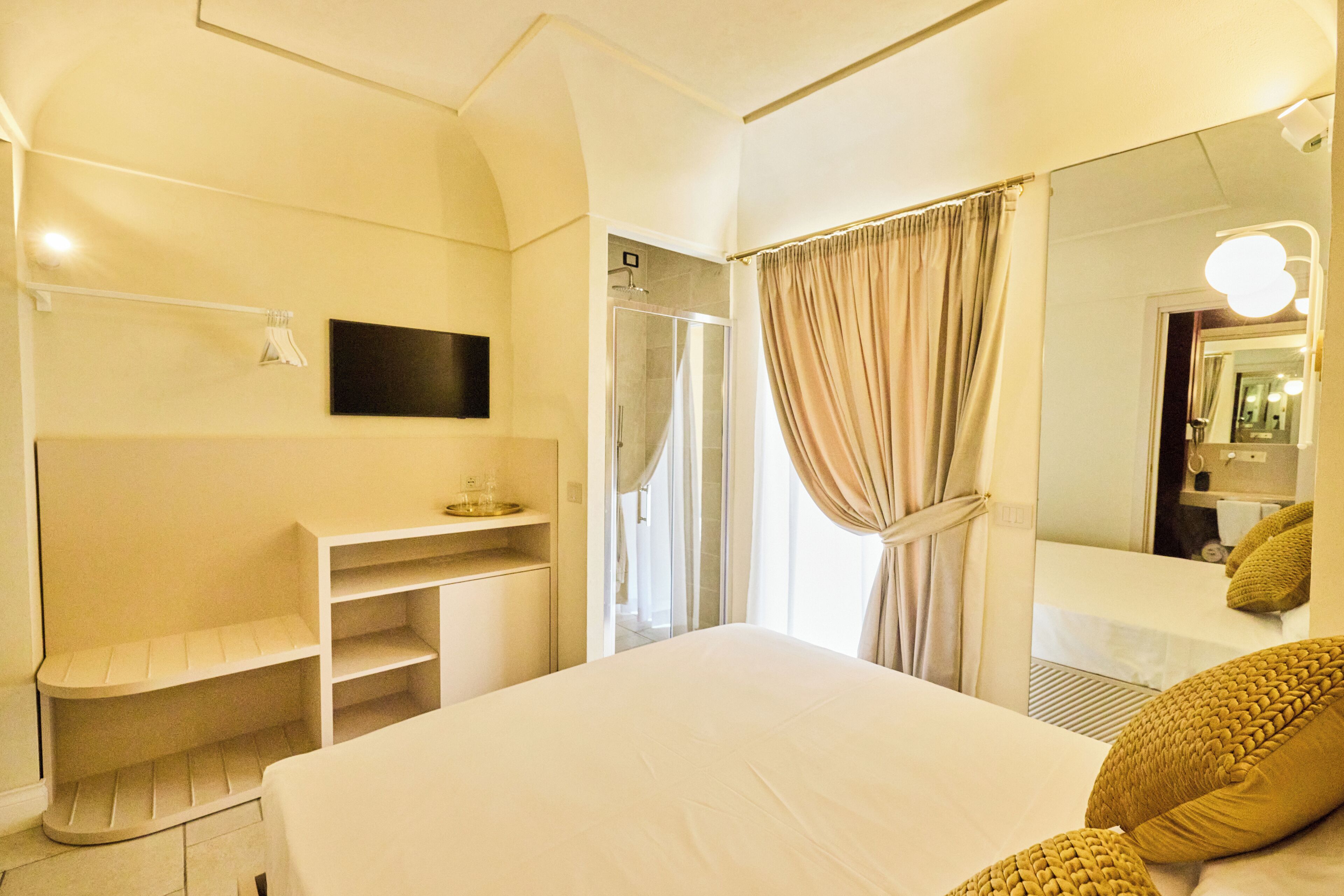 A hotel room with a bed, a vanity with a mirror, a shower, and a TV mounted on the wall.